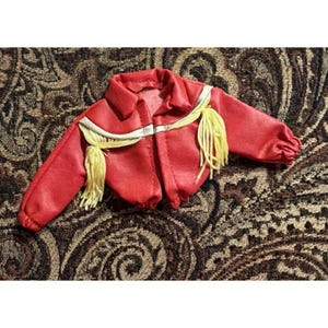 May include: A miniature red jacket featuring a pointed collar, long sleeves with gathered cuffs, and a horizontal yoke across the chest. The garment is decorated with light yellow fringe trim along the front yoke and shoulder seams.
