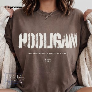 May include: A dark brown t-shirt featuring the white distressed text HOOLIGAN across the chest. Below the main text, it reads MISUNDERSTOOD SINCE DAY ONE. and ESTD 1996. The shirt is worn under a cream-colored cardigan with long sleeves.