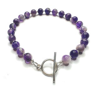 May include: A bracelet made of polished amethyst beads in varying shades of purple. The beads are approximately 0.8 cm in diameter and are strung together. It has a silver toggle clasp. The bracelet is set against a white backdrop.