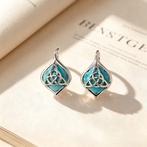Puede incluir: Un par de pendientes de plata con un marco en forma de lágrima y acabado pulido. Cada pendiente contiene una incrustación de piedra turquesa brillante centrada con un intrincado diseño de nudo triquetra de plata, creando un aspecto simétrico.
