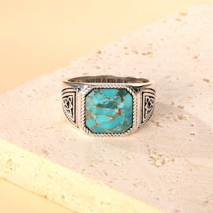 May include: A silver ring with a large, square turquoise stone. The stone has a matrix of gold-colored veins. The ring has a rope-like border around the stone and decorative Celtic knot designs on the sides. The text "IN THE PALM OF HIS HAND" is engraved on the band.