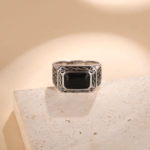 Puede incluir: Anillo de plata con una piedra negra rectangular en el centro. El anillo tiene un diseño ornamentado en los laterales y alrededor de la piedra. Se muestra sobre una superficie texturizada de color blanco roto, sobre un fondo suave y neutro.