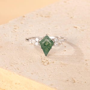 Puede incluir: Un anillo de plata con una gema de ágata musgosa verde en forma de cometa. El anillo está adornado con pequeñas gemas claras a ambos lados de la piedra central. El anillo está sobre una superficie clara y texturizada.