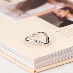 Puede incluir: Un anillo plateado con un diseño curvo único. El anillo está colocado sobre un libro de color claro, con un fondo borroso. La forma del anillo es una curva continua y fluida, creando una estética moderna y minimalista.