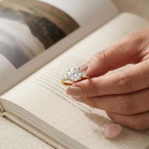 Puede incluir: Un anillo de oro con una gema grande, redonda y transparente en el centro, flanqueada por dos gemas más pequeñas. El anillo se sostiene contra el telón de fondo de un libro abierto. Una pequeña piedra rosa pulida está en la esquina inferior derecha.