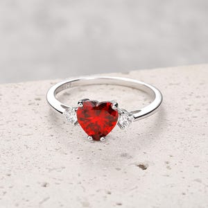 Könnte beinhalten: Ein silberner Ring mit einem herzförmigen, tiefroten Edelstein als Mittelpunkt. Zwei kleinere, klare Edelsteine flankieren das Herz. Der Ring ist vor einem neutralen Hintergrund platziert, der das Design des Schmucks hervorhebt.