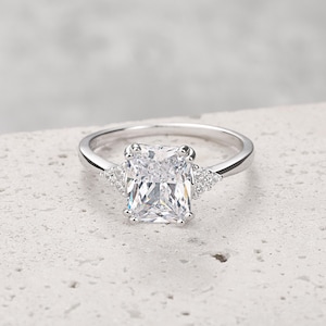 Op de afbeelding: Een zilveren ring met een grote, rechthoekige, heldere edelsteen als middelpunt. Twee kleinere, driehoekige clusters van heldere stenen flankeren de hoofdsteen. De ring ligt op een getextureerd, lichtgekleurd oppervlak.