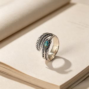 Puede incluir: Un anillo abierto de tono plateado con forma de pluma y líneas grabadas detalladas. El anillo presenta una pequeña piedra ovalada de color turquesa engastada en el centro del diseño de pluma, creando un contraste marcado con el acabado metálico.