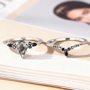 Puede incluir: Dos anillos de plata. Uno presenta una piedra en forma de pera, gris y transparente, con inclusiones negras y piedras de acento negras. El otro es una banda de plata con piedras negras en forma de marquesa y redondas.