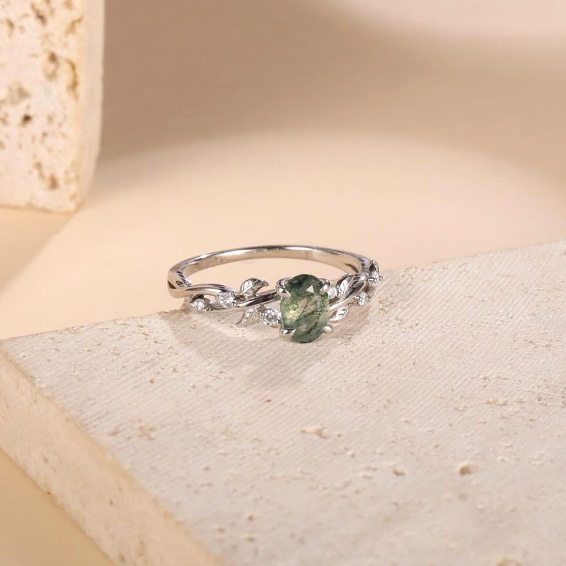 May include: A silver-toned ring featuring an oval-shaped moss agate center stone. The band is designed with intricate metal leaf motifs and small, clear, round-cut accent stones embedded along the sides of the setting, creating a botanical look.