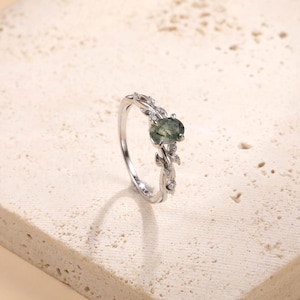 May include: A silver-toned ring featuring an oval-shaped moss agate center stone. The band is designed with a metallic leaf motif that extends along the sides, accented with small, clear, round-cut stones embedded within the metal foliage.