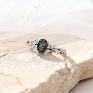 Puede incluir: Un anillo de plata con una piedra preciosa ovalada de color gris oscuro. El anillo presenta un delicado diseño de vid con pequeñas piedras de acento transparentes. El anillo se muestra sobre una superficie clara y texturizada.