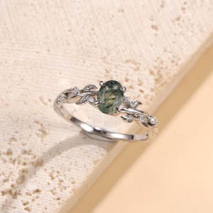 May include: A silver-toned ring featuring an oval-cut moss agate center stone. The band is designed with a delicate leaf motif on both sides, accented with small, clear round stones embedded along the metal foliage, creating a nature-inspired look.