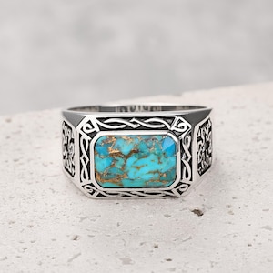 Puede incluir: Un anillo de plata con una piedra turquesa rectangular. El anillo tiene un diseño ornamentado alrededor de la piedra y en los lados. La piedra turquesa es de color azul-verdoso con motas doradas.