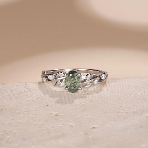 May include: A silver ring with an oval, green gemstone at the centre. The ring features a leaf and vine design with small, clear accent stones. The ring is set against a neutral background.