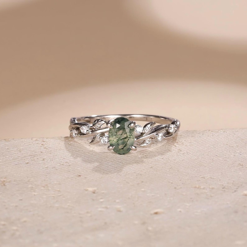 May include: A silver-toned ring featuring an oval-cut moss agate center stone with green inclusions. The band is designed with intricate metal leaf motifs and small, clear round-cut accent stones embedded along the sides of the setting.