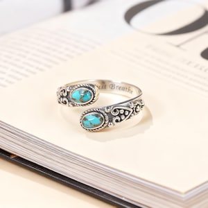 Può includere: Un anello color argento con due pietre ovali turchesi. L'anello dal design aperto presenta decorazioni a volute e le parole "Just Breathe" incise sulla fascia. L'anello è esposto su un libro aperto.