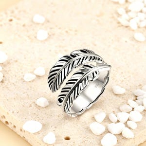 Pode incluir: Um anel prateado com um design de pena. O anel apresenta três faixas sobrepostas em forma de pena com linhas detalhadas, criando um efeito em camadas. O anel está sobre um fundo neutro com pequenas pedras brancas.