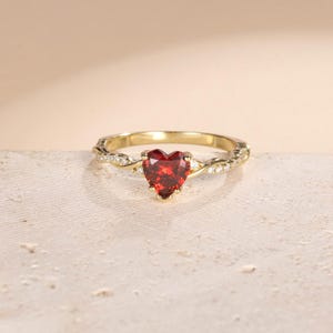 Op de afbeelding: Een gouden ring met een hartvormige, rode edelsteen. De band heeft een gedraaid ontwerp met kleine, heldere accentstenen. De ring staat tegen een neutrale achtergrond, wat het elegante ontwerp benadrukt.