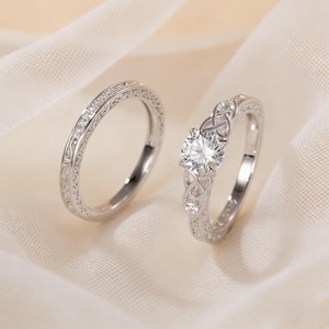 Puede incluir: Dos anillos de plata. Uno es una banda con un patrón detallado y pequeñas piedras. El otro anillo tiene una piedra grande, redonda y transparente y un diseño de nudo celta. Ambos anillos están sobre un fondo blanco.