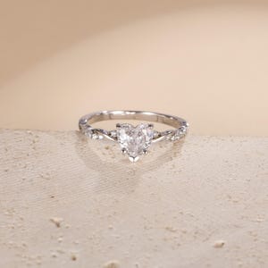 May include: A silver ring with a heart-shaped diamond center stone. The band features a twisted design with small accent stones. The ring is set against a neutral background.