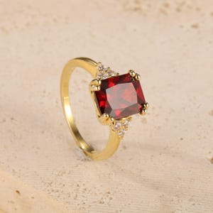 Puede incluir: Un anillo de oro con una gran piedra preciosa rectangular de color rojo intenso. El anillo tiene pequeñas piedras de acento claras a cada lado de la piedra principal. La banda es de oro pulido simple. Una joya clásica.