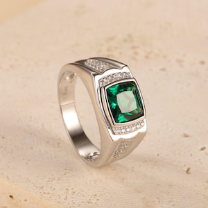 Puede incluir: Anillo plateado con una gema cuadrada de color verde esmeralda. El anillo tiene una cara rectangular con pequeñas piedras claras. La banda es lisa y pulida, con un diseño clásico.