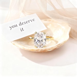 May include: A gold ring with a large, clear, rectangular-cut gemstone. The ring is displayed on a seashell with a card that reads "you deserve it". The background includes a soft, draped fabric.
