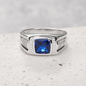 Puede incluir: Un anillo plateado con una gran piedra preciosa cuadrada de color azul oscuro en el centro. El anillo tiene una cara rectangular con piedras más pequeñas y claras a cada lado. La banda es pulida y reflectante, sobre un fondo neutro.