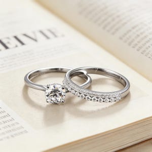 Puede incluir: Dos anillos de plata sobre una página de libro. Uno presenta una gema grande, redonda y transparente. El otro tiene una banda de pequeñas gemas transparentes. Los anillos están sobre un fondo neutro.