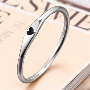 Puede incluir: Un anillo plateado con un pequeño diseño de corazón negro. El anillo es liso y brillante, con la marca "S925" visible en la banda. Una pieza de joyería sencilla y elegante.