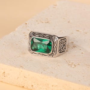 Puede incluir: Un anillo plateado con una gran piedra preciosa rectangular de color verde esmeralda. El anillo presenta detalles ornamentados alrededor de la piedra preciosa y en los laterales, con un diseño cuadrado. El anillo está sobre una superficie de color neutro.