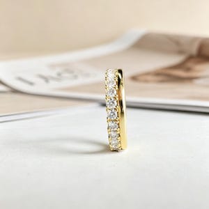 Puede incluir: Un anillo dorado con una fila de pequeñas piedras preciosas transparentes. El anillo se muestra verticalmente, destacando el diseño y el brillo de las piedras. El fondo está borroso, resaltando los detalles del anillo.