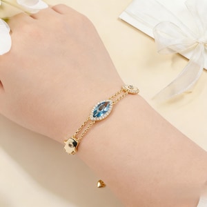 Puede incluir: Pulsera dorada con una gema azul ovalada en el centro, rodeada de pequeñas piedras claras. La pulsera tiene un diseño de cadena fina con detalles florales. Una joya elegante para cualquier ocasión.