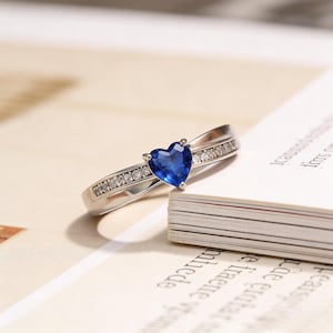 May include: A silver ring with a heart-shaped, deep blue gemstone at the center. The band is adorned with small, clear gemstones. The ring is set against a backdrop of a book.