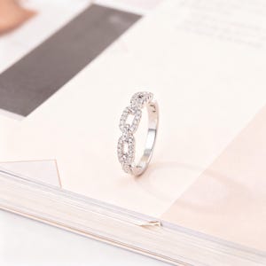 Puede incluir: Un anillo plateado con un diseño de eslabones de cadena, incrustado con pequeñas piedras preciosas transparentes. El anillo se presenta sobre un fondo suave y neutro, resaltando sus intrincados detalles y brillo. Una joya.