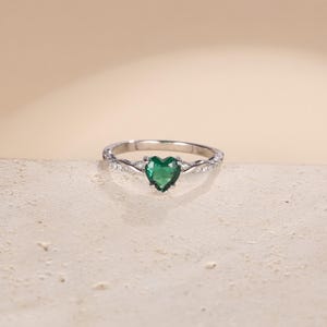 Puede incluir: Un anillo de plata con una piedra preciosa verde esmeralda en forma de corazón. La banda tiene un diseño retorcido con pequeñas piedras de acento claras. El anillo se muestra sobre una superficie texturizada neutra, resaltando su elegante diseño.