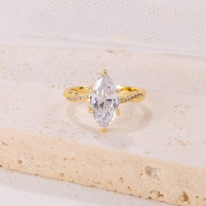 Puede incluir: Un anillo dorado con una gran gema transparente de talla marquesa. La banda tiene un diseño retorcido con pequeños detalles brillantes. El anillo se muestra sobre un fondo neutro y texturizado.