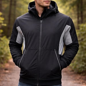 May include: A black hooded jacket with gray shoulder panels and a front zipper. The jacket has two front pockets with white trim and is worn with blue jeans. The background is a blurred forest scene.