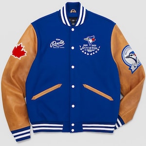 Puede incluir: Chaqueta universitaria azul y marrón con puños y cuello a rayas blancas y azules. La chaqueta presenta un parche de hoja de arce, logotipos de equipos y el texto "Roots" y "Har Bast Division".