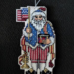 May include: A cross-stitch ornament featuring a figure with a white beard wearing a blue jacket with white stars, red and white striped trousers, and a hat. The figure holds a small American flag, a drum, and a gold trumpet, suspended by a silver string.