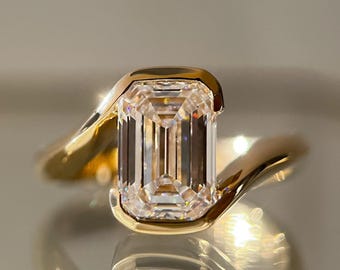 May include: A gold ring featuring a large, rectangular, clear gemstone. The gemstone is set in a gold band that curves around the finger. The ring is highly reflective, with light reflecting off the gemstone and the gold.