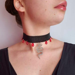 May include: A black beaded choker necklace featuring a series of small, dangling red teardrop-shaped charms along the bottom edge. A silver-toned metal pendant shaped like a coiled snake with a crescent moon detail hangs from the center.