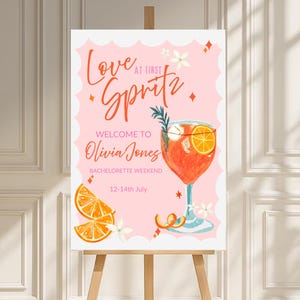 May include: A pink sign on a wooden easel, featuring the words "Love at First Spritz" in orange script. The sign welcomes guests to Olivia Jones' bachelorette weekend, with a watercolour illustration of a cocktail and orange slices.