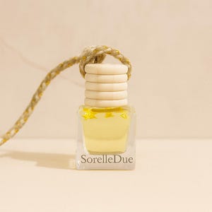 May include: A small, clear glass bottle containing a pale yellow liquid and star-shaped decorations. It has a wooden cap and a woven gold-coloured string for hanging. The text "SorelleDue" is printed on the front.