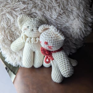 May include: Two handmade crochet bunny dolls. One bunny has a red bow and red accents on its face. The other bunny has a ruffled collar. Both are cream-colored and sit in front of a fluffy, white pillow.
