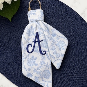 May include: A fabric keychain featuring a light blue and white floral pattern with a dark blue embroidered letter A. The fabric is looped through a gold-toned metal ring, creating a soft, folded appearance with two pointed ends hanging down.