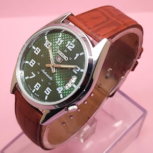Könnte beinhalten: Eine Seiko Automatik-Armbanduhr mit grün gemustertem Zifferblatt und silbernem Gehäuse. Die Uhr hat ein braunes Lederarmband. Das Zifferblatt zeigt die Zeit mit weißen Ziffern und Zeigern an. Hergestellt in Japan.