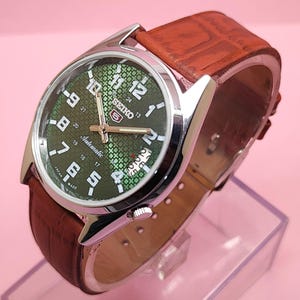 May include: A Seiko automatic wristwatch with a green patterned face and silver case. The watch has a brown leather band. The watch face displays the time with white numerals and hands. The watch is made in Japan.
