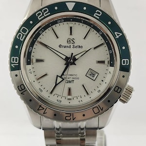 May include: A stainless steel Grand Seiko automatic watch with a white dial, green bezel, and silver band. The watch face displays the date, hour markers, and the text "GS Grand Seiko AUTOMATIC - BEAT 36000 GMT".